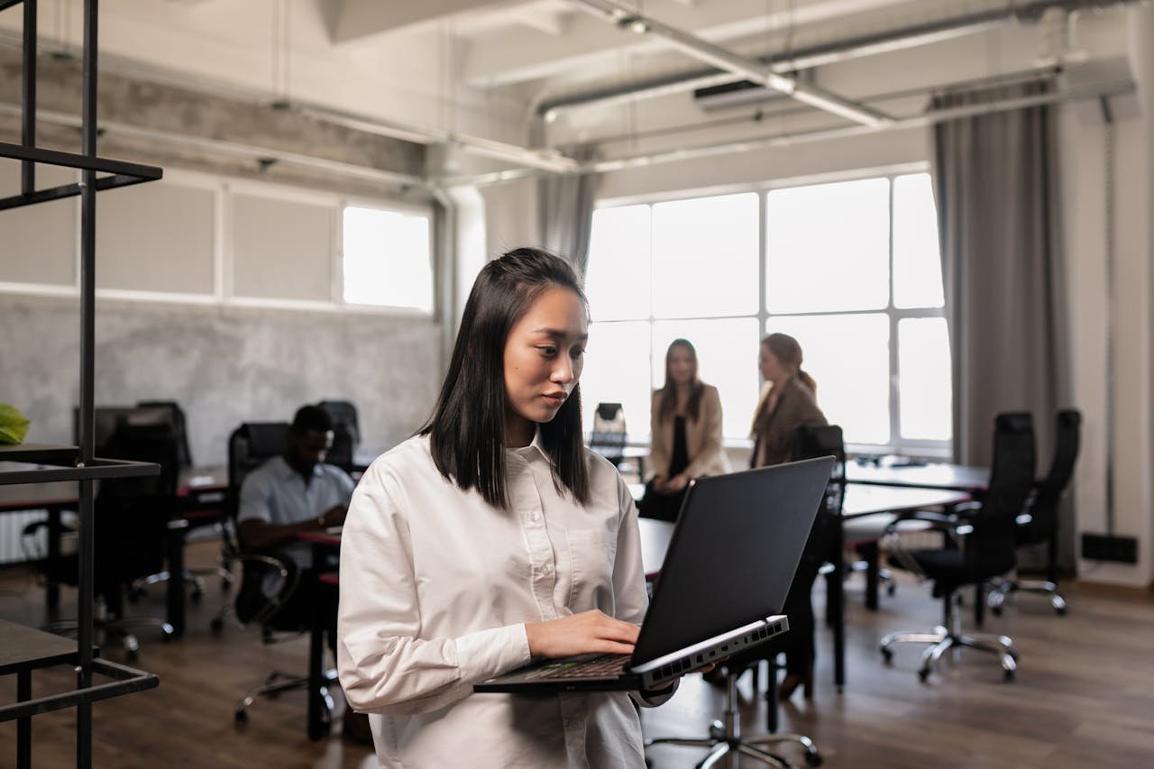 A focused woman using a laptop in a spacious, well-lit office environment with colleagues nearby.